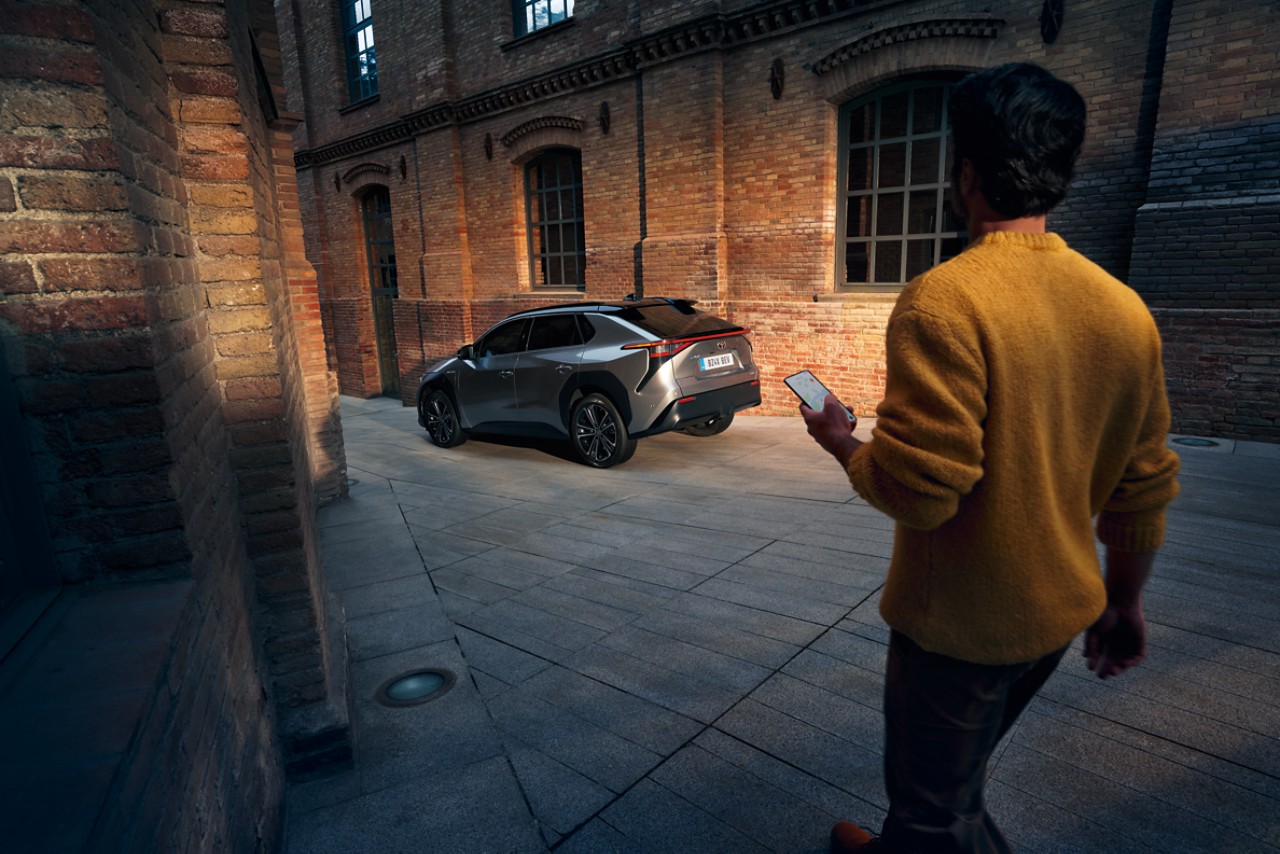 A man sits in the driver's seat of a Toyota car holding his smartphone. The phone displays the TOYOTA MyT app logo.